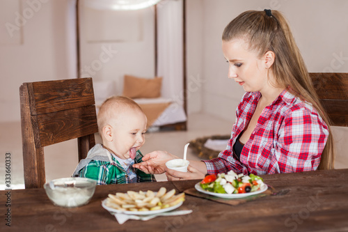  Beautiful girl in a red checkered shirt sits at a wooden brown table and feeds the child. A one-year-old boy in a green shirt choked on food and stuck out his tongue.