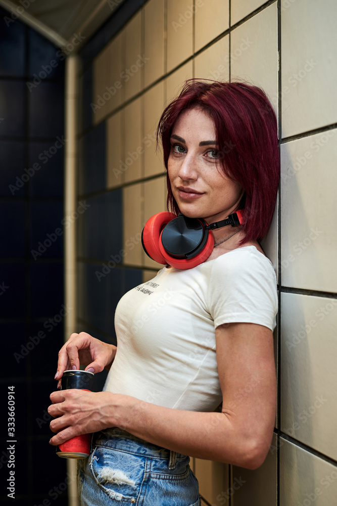 Young woman with burgundy pink maroon hair, in white top and light blue  jeans, leaning on