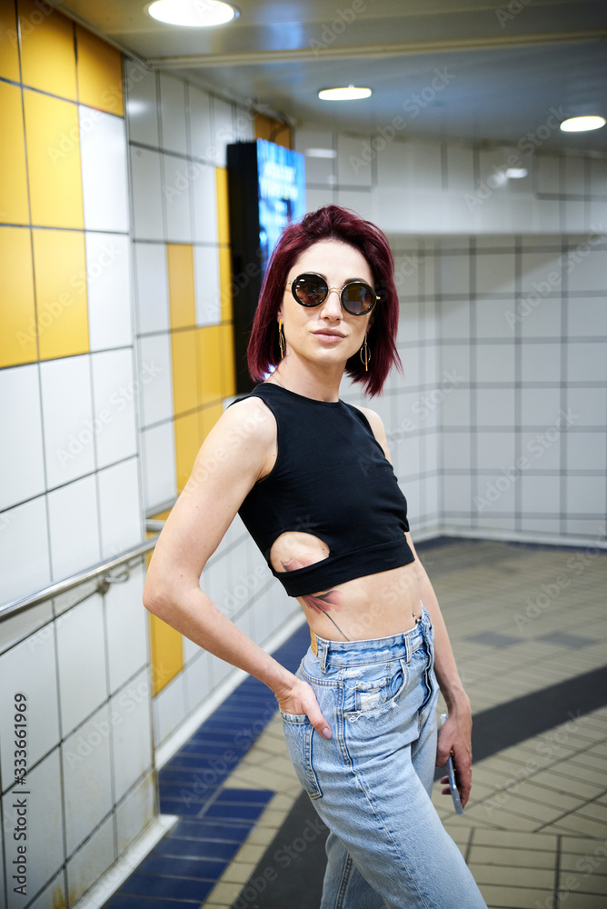 Young woman with burgundy red maroon short hair, wearing black top, light blue jeans, standing in subway station, Three-quarter portrait of pretty girl in black sunglasses at metro station Young woman with burgundy red maroon short hair, wearing black top, light blue jeans, standing in subway station, Three-quarter portrait of pretty girl in black sunglasses at metro station