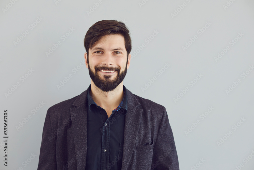 Positive bearded guy in a jacket smiling looks at the camera on a gray background. - obrazy, fototapety, plakaty 