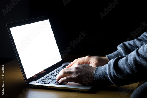 Man working on laptop with white screen glowing in the dark on a wooden desk from the side