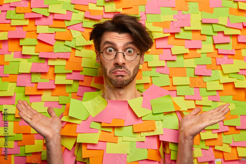 What can I say? Confused man shrugs shoulders and faces dilemma, keeps hands raised in palms, has surprised expression, wears round transparent glasses, poses against colorful stickers background