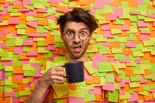 Taking coffee break. Embarrassed man smirks face, looks puzzled as has much work in office, holds cup of tea, wears transparent glasses, poses against colorful stickers, dissatisfied with something photo