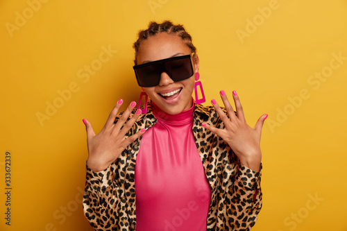 Photo of cheerful stylish woman boasts her new manicure, smiles broadly, wears trendy sunglasses and leopard clothes, isolated on yellow background. Glamour dark skinned lady in fashionable outfit