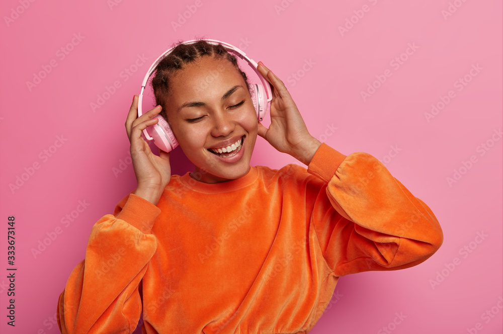 Modern life, fun and entertainment concept. Happy ethnic girl listens song or cool track in stereo headphones, being on positive tune, enjoys best hits from playlist, closes eyes isolated on pink wall Modern life, fun and entertainment concept. Happy ethnic girl listens song or cool track in stereo headphones, being on positive tune, enjoys best hits from playlist, closes eyes isolated on pink wall
