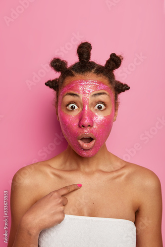 Image of shocked ethnic woman applies rosy cosmetic mask on face  removes blackheads or pores  points at herself  stares with bugged eyes  hears unexpected advice from beautician  has knots on head