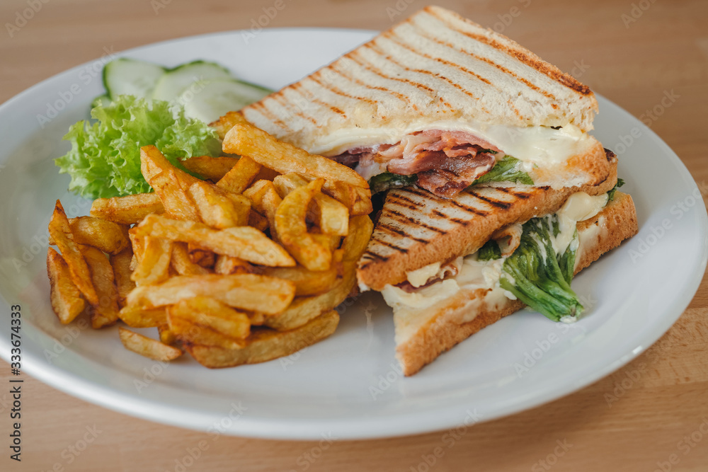 Bacon club sandwich with fries and vegetables on a white plate,on a wooden background. Meal time. Bacon club sandwich with fries and vegetables on a white plate,on a wooden background. Meal time.
