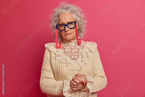 Horizontal shot of serious wise elderly woman has curly grey hair, wrinkled face, keeps hands together, makes photo for family album, wears glasses, poses over pink background. Mature female portrait