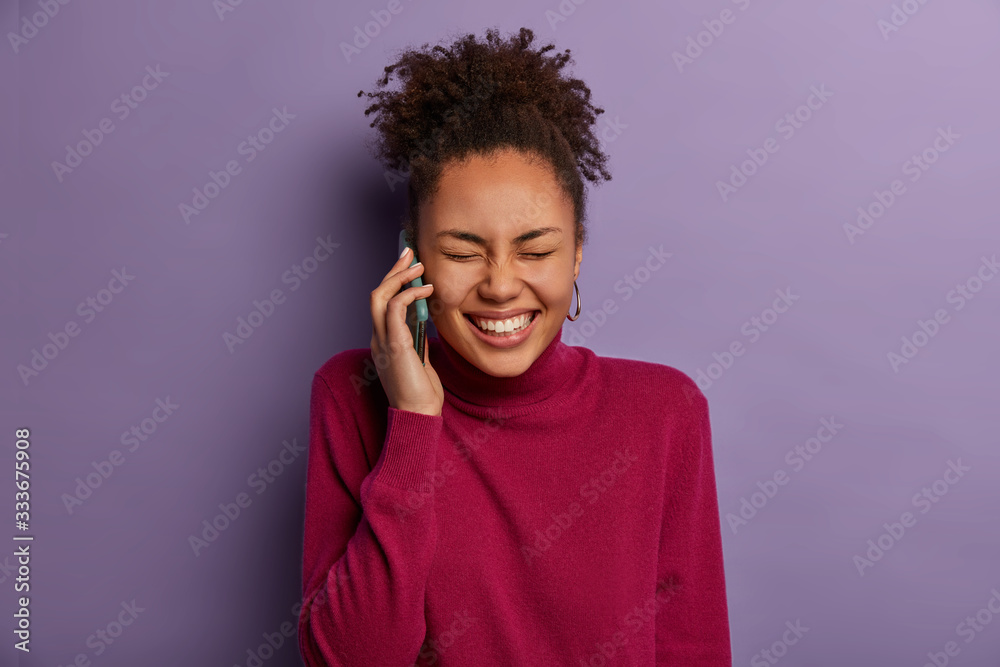 Photo of overjoyed ethnic woman has funny talk, keeps modern mobile phone near ear, smiles broadly, cannot stop laughing, has curly combed hair, dressed casually, isolated on purple background Photo of overjoyed ethnic woman has funny talk, keeps modern mobile phone near ear, smiles broadly, cannot stop laughing, has curly combed hair, dressed casually, isolated on purple background