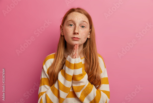 I am sorry! Pleading ginger girl keeps palms pressed together, begs parents for forgiveness, asks to give one more chance to improve, looks with sad expression, dressed in casual loose jumper