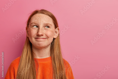 Funny freckled redhead girl looks happily aside, has tricky plan, smiles with cunny expression, dressed casually, isolated on pink background, copy space for your information. Happy child plays alone