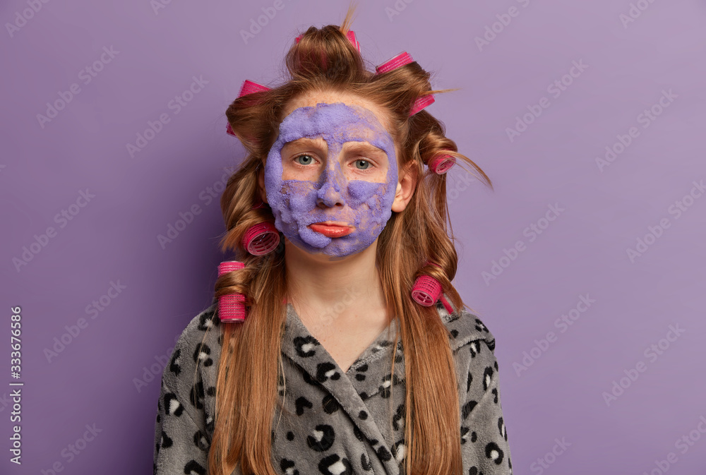 Offended displeased ginger girl looks upset, feels bored because of beauty treatments, applies face mask and hair curlers, prepares for birthday party, wants to look perfect, wears soft bathrobe