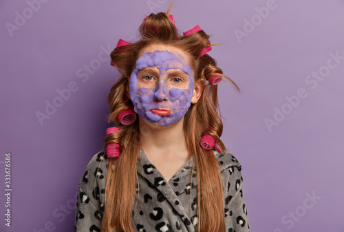 Offended displeased ginger girl looks upset, feels bored because of beauty treatments, applies face mask and hair curlers, prepares for birthday party, wants to look perfect, wears soft bathrobe