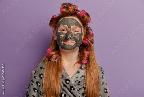 Horizontal shot of beautiful smiling girl appplies face mask, enjoys cosmetic treatment at home, wants to look nice, wears hair curlers for making hairstyle, robe, isolated on purple background