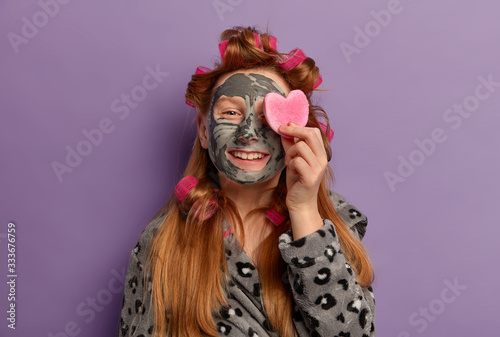 Natural beauty small girl applies clay mask  covers eye with heart shaped sponge  feels happy  makes hairstyle with curlers  enjoys young skin  uses cosmetic products for kids  wears bathrobe