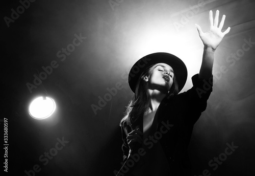 Obraz na plátně Beautiful braless actress girl, wearing an unbuttoned blazer and a hat, poses and dances in the warm light rays of scenic illuminators in the theatrical fog
