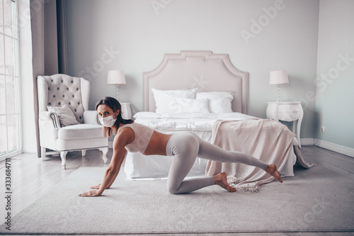 Home fitness. Young fit slim woman in protection face mask doing morning work-out yoga exercises in bedroom during self isolation quarantine. COVID-19 concept to promote stay safe home save lives