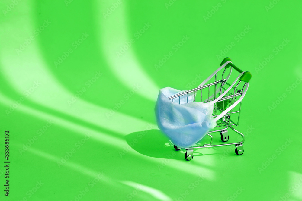 An empty shopping cart with a face mask on green background. Consumer buying panic about coronavirus covid-19 concept, home quarantine. Home delivery