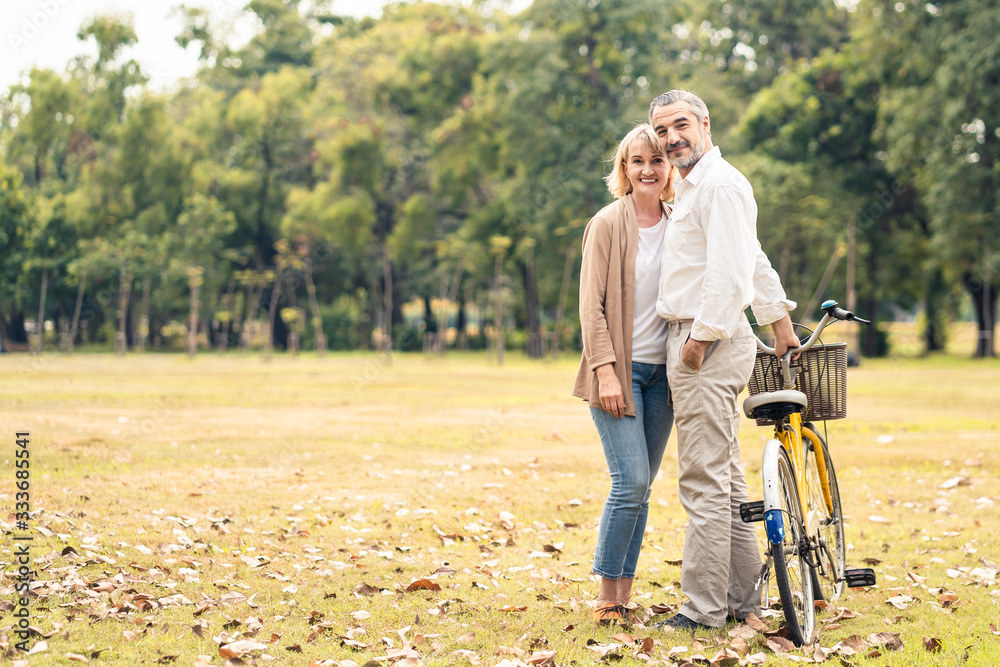 Caucasian senior elder couple stand & hold bicycle in park. Mature are happy and enjoy with slow life. Two people delight activity, look smile at camera together. Retirement family lifestyle concept.