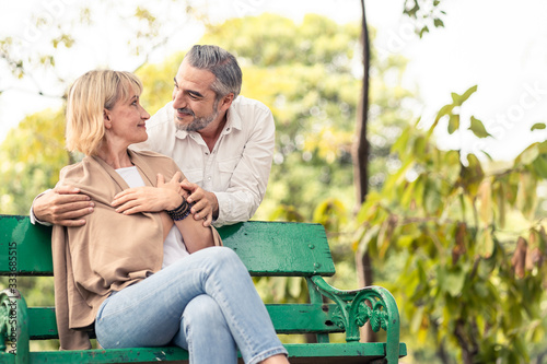 Caucasian senior elder couple sit on bench in park. Mature are happy and enjoy with slow life. Old man love, hug woman from behind. They look and smile together. Retirement family lifestyle concept. © Kawee