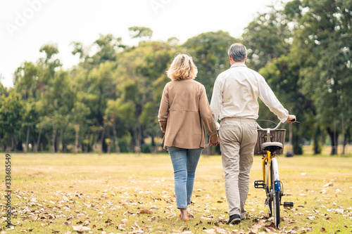 Caucasian senior elder couple love to hold hand and carry bicycle walk in park. Mature are happy and enjoy with slow life. Two people delight activity together. Retirement family lifestyle concept.