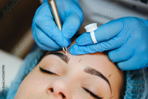 Permanent make-up for eyebrows of beautiful brunette woman in beauty salon. Closeup beautician in gloves doing microblading for eyebrow. photo
