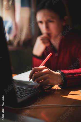 Young caucasian girl in red sweater with confused look on her face looking at computer while studying online at home with pen in hand near the window