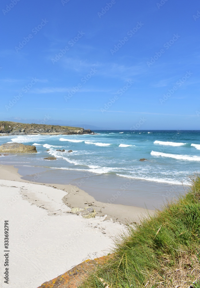 Wild beach with waves, white sand and cliff with blue sky. Viveiro, Lugo, Galicia, Spain.