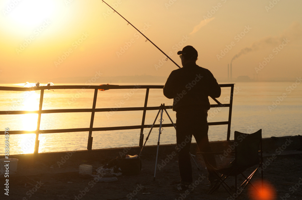 Red sunset on the still sea, black silhouette of a man fishing with a rod, industrial view far in front of him
