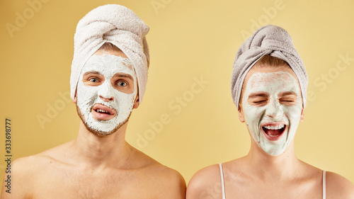 Young friends guy and girl with funny expressive emotions, towel on head and face mask isolated on a yellow background, skin care concept