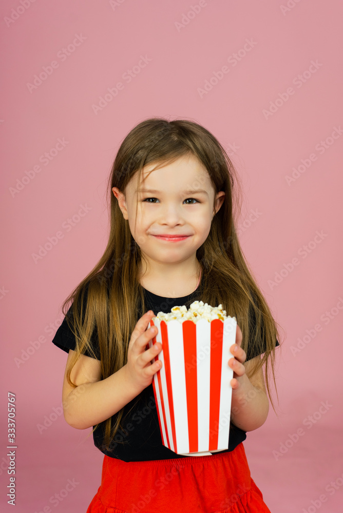 Happy cute little girl holding popcorn in her hands and looking at the camera on a pink background. The concept of watching a movie in a movie theater or at home. Copyspace Happy cute little girl holding popcorn in her hands and looking at the camera on a pink background. The concept of watching a movie in a movie theater or at home. Copyspace