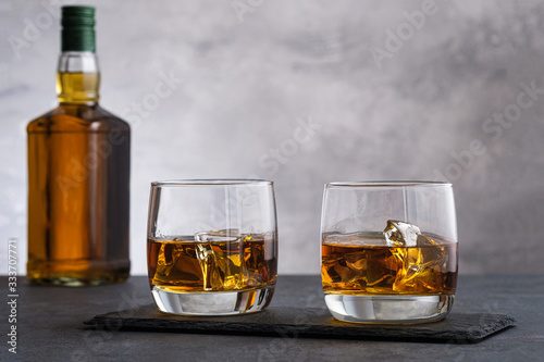 Two glass with whiskey and ice cubes on background bottle