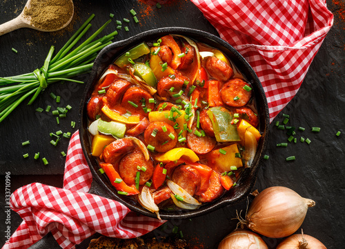 Stewed pepper and sausage with tomatoes in a cast iron skillet on a black background, top view, close up. Traditional Hungarian stew called lecho  photo