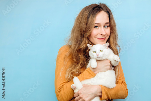 A young girl is holding a white cat in her arms. Portrait of a curly-haired blonde girl on a blue background. The concept of animal protection. Take the cat from the shelter.