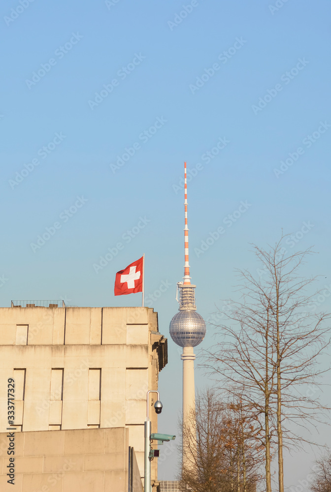 Red and white Swiss flag waving over the embassy of Switzerland with the sunlit Berlin TV tower in the background on a cloudless sunny day.