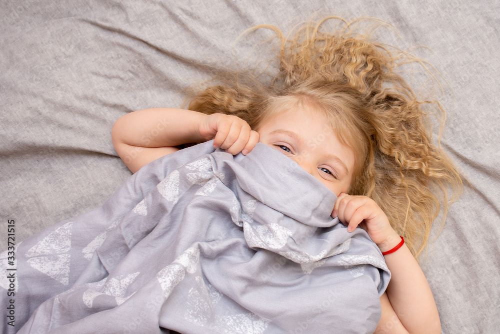 Portrait of a blue-eyed little girl with blond curly hair waking up in the morning on a gray bed laughing merrily. Portrait of a blue-eyed little girl with blond curly hair waking up in the morning on a gray bed laughing merrily.