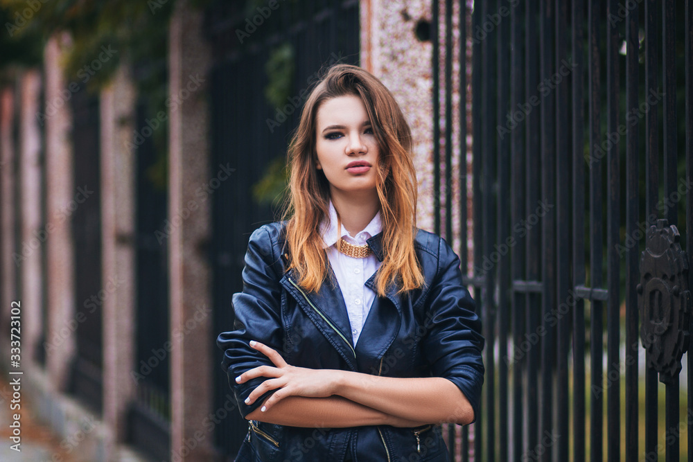 Young beautiful fashionable European woman in a black leather jacket on a background of a dark metal fence.