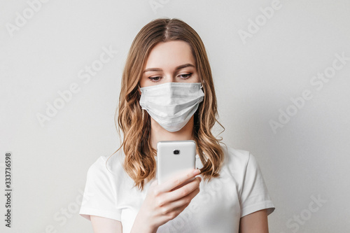 Young girl in a medical mask calls on the phone and looking at a cell phone isolated over light grey background, ordering goods, eating online, quarantine, isolation, coronovirus, copy space © Evgenia