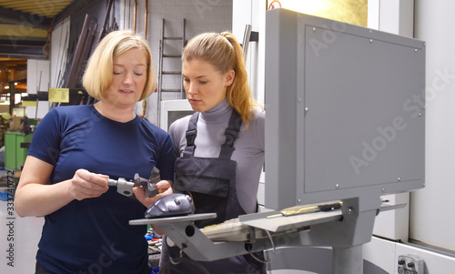 A young trainee receives tuition from her trainer. Both women work as computerized  numerical control engineers. They are seen  together at their high tech place of work.