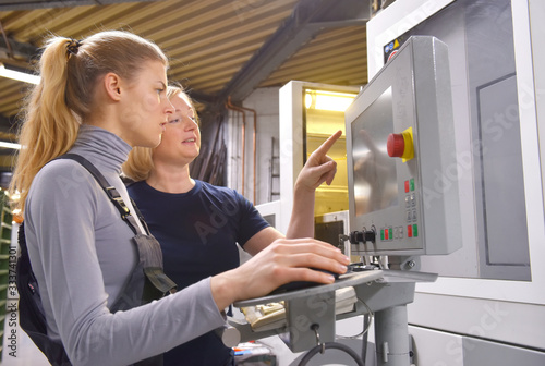 A young trainee receives tuition from her trainer. Both women work as computerized  numerical control engineers. They are seen  together at their high tech place of work.