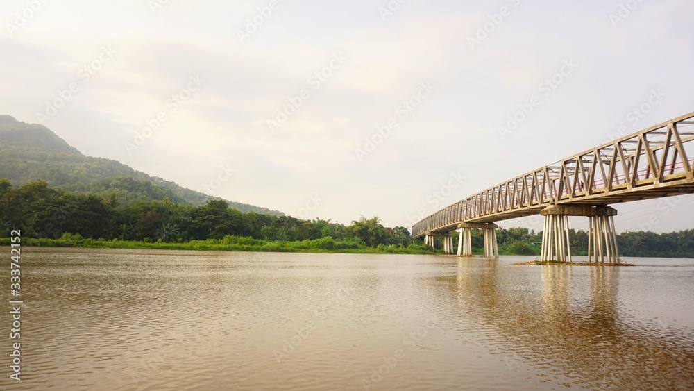 Obraz premium A view on the river bank and a long bridge over the river. Visible mountains across the river.