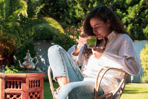 Arabian young woman with ethnic features smelling peppermint arabic tea at her garden. Moroccan female preparing traditional arab tea at home. Arabian culture and traditions. Muslim lifestyle.