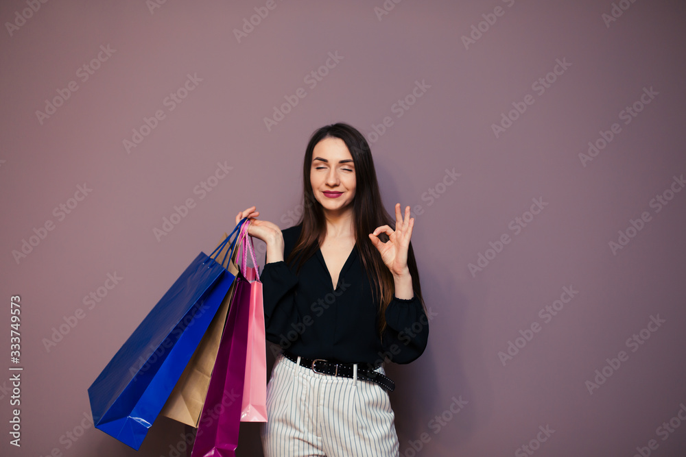 Happy young woman wearing blouse standing isolated on purple background carrying shopping bags showing okay gestures Happy young woman wearing blouse standing isolated on purple background carrying shopping bags showing okay gestures