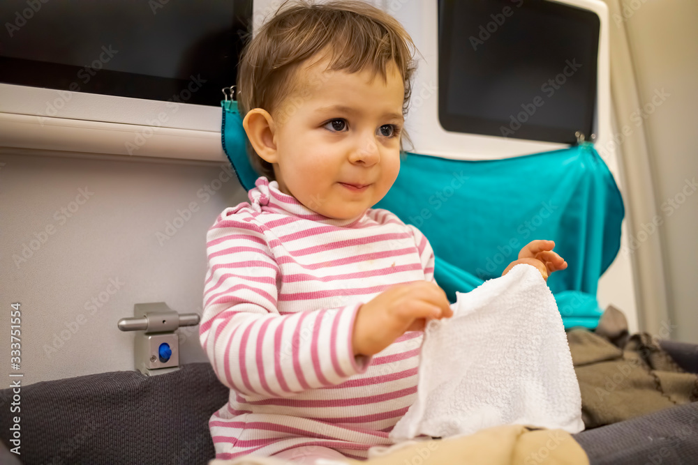 happy little cute baby boy sitting in the baby bassinet of the airplane, smiling and holding a wet, hot towel for washing hands and face after sleeping on a long night flight. happy little cute baby boy sitting in the baby bassinet of the airplane, smiling and holding a wet, hot towel for washing hands and face after sleeping on a long night flight.