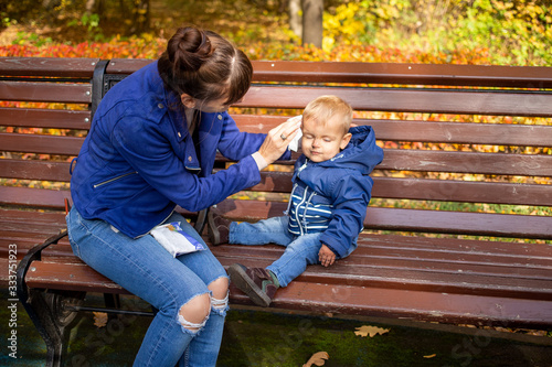 young beautiful mom in blue clothes on a bench in the park wipes the baby’s dirty face with a napkin. baby is in blue clothes. close-up, soft focus, on the background of trees in blur