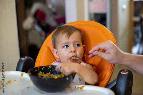 hand in a plate and eats from the spoon held by the parent. close-up, front view, soft focus, blur background