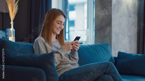 Happy beautiful young woman lying on the sofa and using the phone, typing, scrolling, chatting and smiling at modern room.