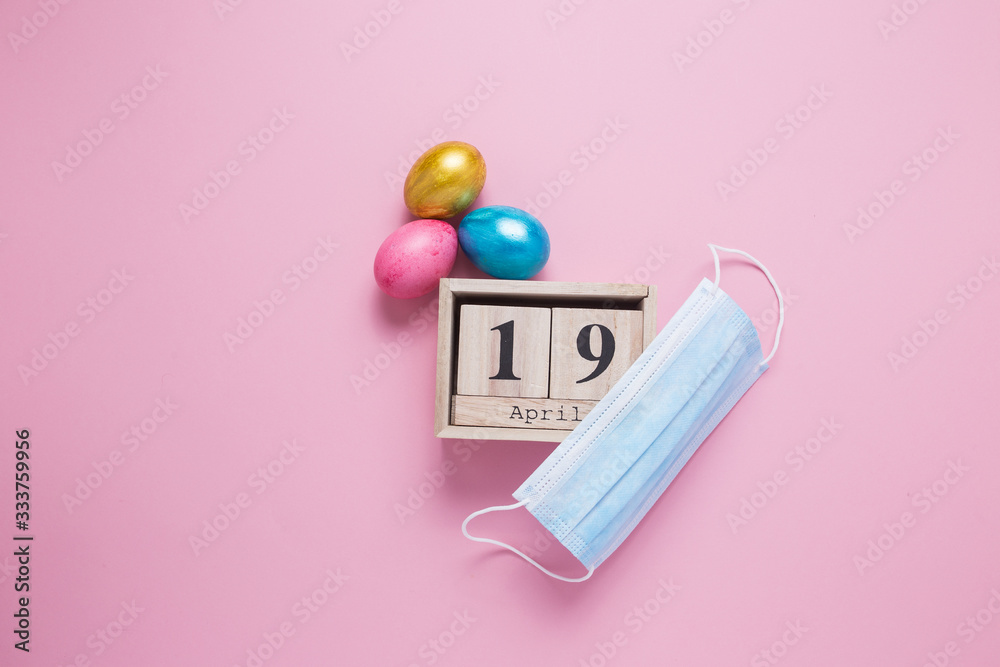 Wooden cube with the date of the celebration of Easter on April 19. Medical mask ban on mass celebrations. Easter eggs on a pink background. COVID-19. Wooden cube with the date of the celebration of Easter on April 19. Medical mask ban on mass celebrations. Easter eggs on a pink background. COVID-19.