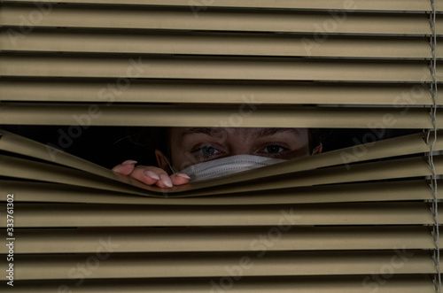 Young woman in medical mask looks out the window through the blinds