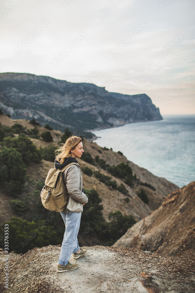 Happy young traveler woman walking and enjoying amazing coastline mountain view. With beige backpack, wearing in jacket and jeans.